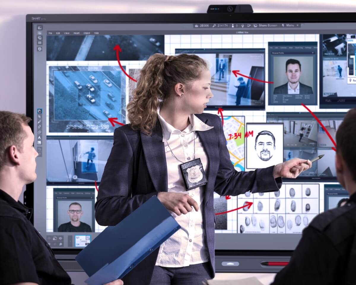 A female investigator points at a SMART interactive display showing multiple surveillance feeds, including car surveillance and suspect tracking, during a strategic discussion.