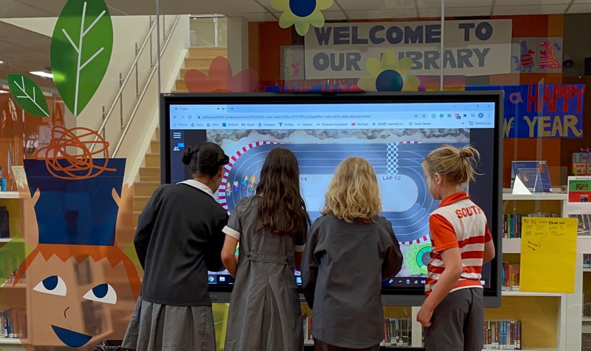 A group of students actively engages with a SMART Board in a library setting, collaboratively interacting with an educational program displayed on the screen, illustrating the multi-user experience feature of the device.