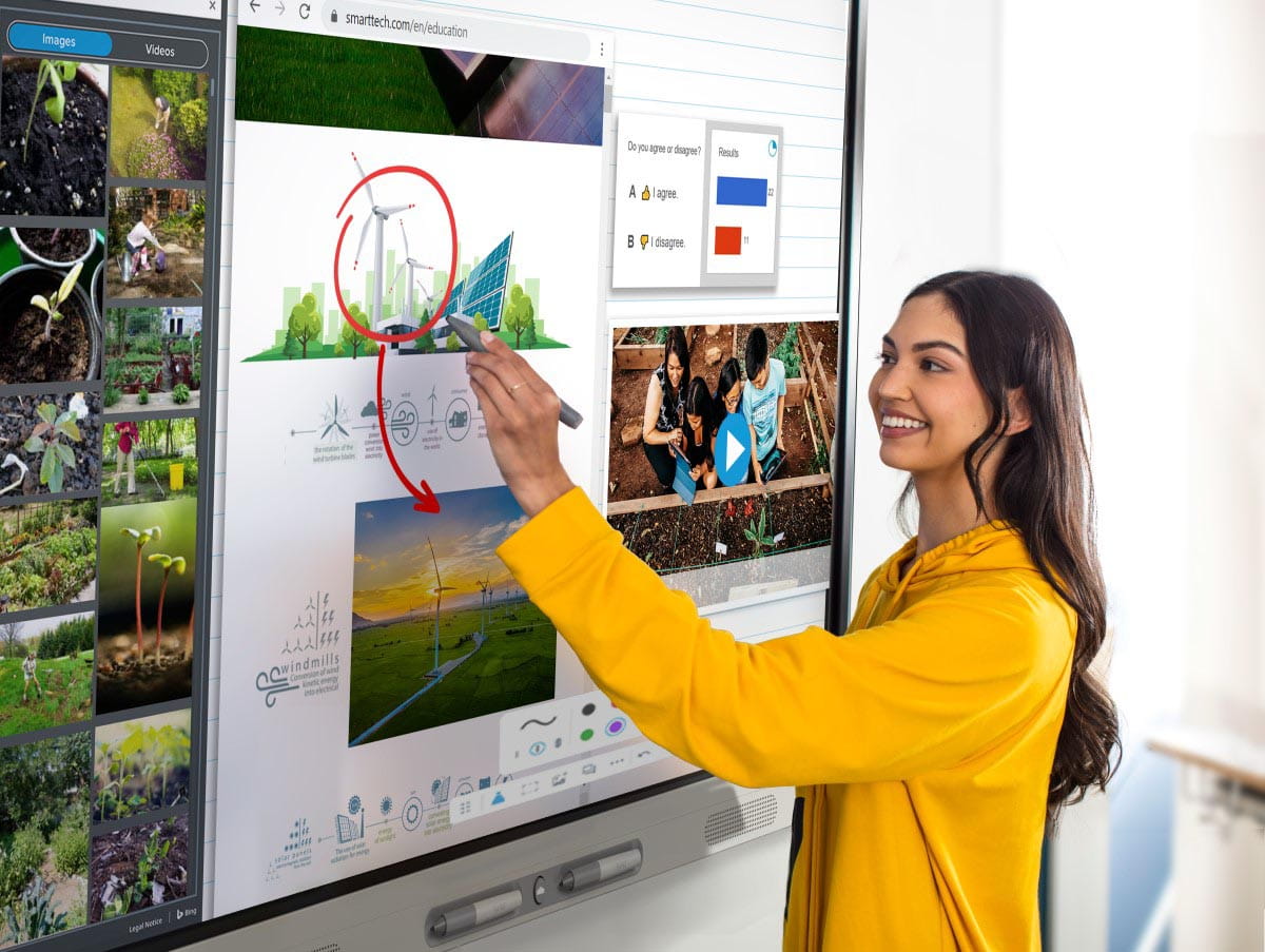 A teacher in a yellow sweater is interacting with a SMART Board MX series display, circling a diagram of wind turbines on a lesson about renewable energy, with a web browser open in the background displaying related images and videos.