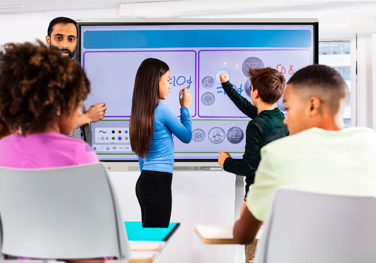 In a brightly lit classroom, a teacher and two students interact with a SMART Board, with the students using digital manipulatives on the screen to solve a math problem involving coins.