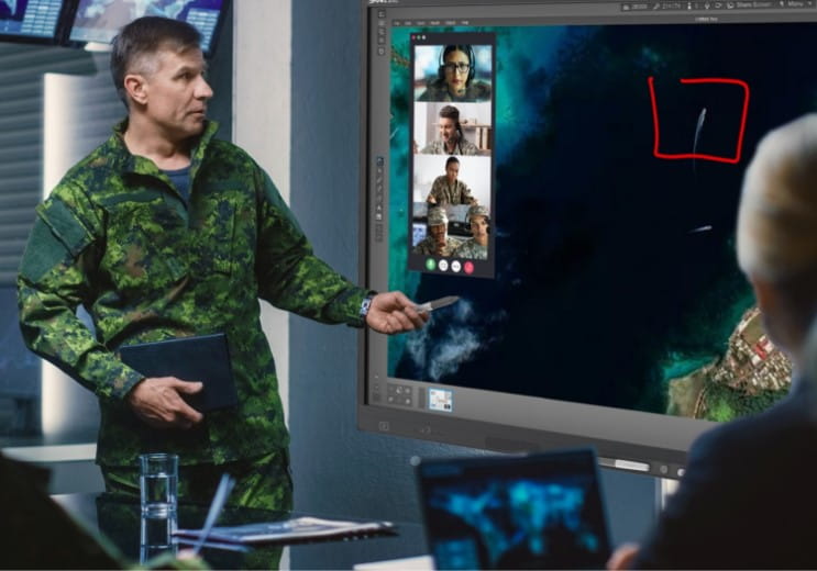 A male military officer in camouflage uniform standing next to a SMART interactive display, pointing to a satellite image of a geographical area with marked locations.