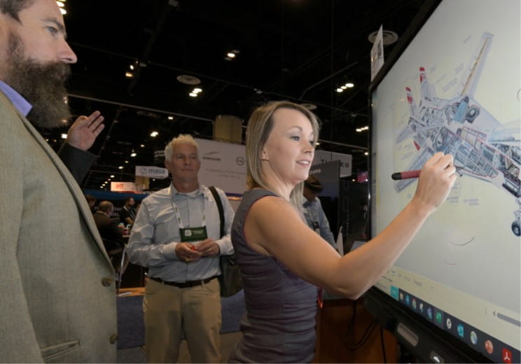 A woman in business attire using SMART Ink to annotate over a digital image of an aircraft on a SMART interactive display at a technology expo.