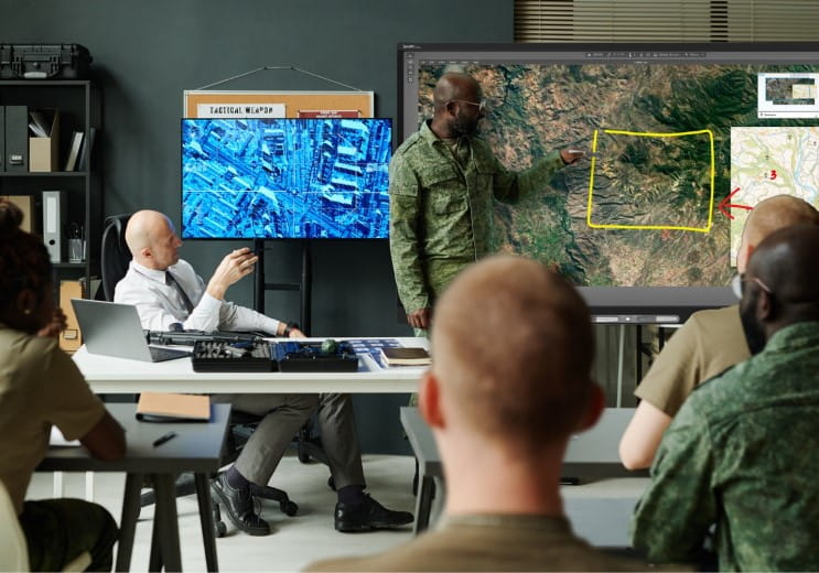 A strategic meeting in a military briefing room with multiple personnel, focusing on a senior officer pointing to a specific area on a satellite image displayed on a SMART interactive display.