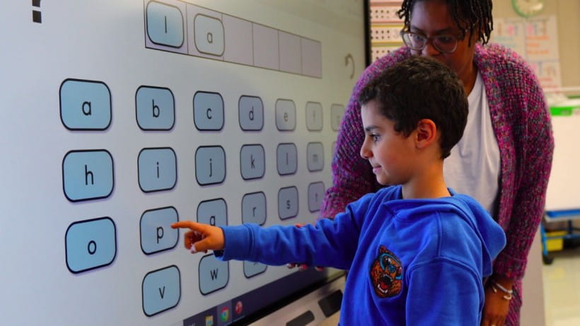 Elementary student interacting with an alphabetical keyboard on a SMART board under the guidance of his teacher in a classroom.