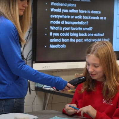 A teacher holding a mic to a student sharing her thoughts with the entire classroom with the help of audio enhancements, in front of a SMART Board.